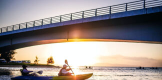 tandem kayakers pass under a bridge while paddling in urban waters