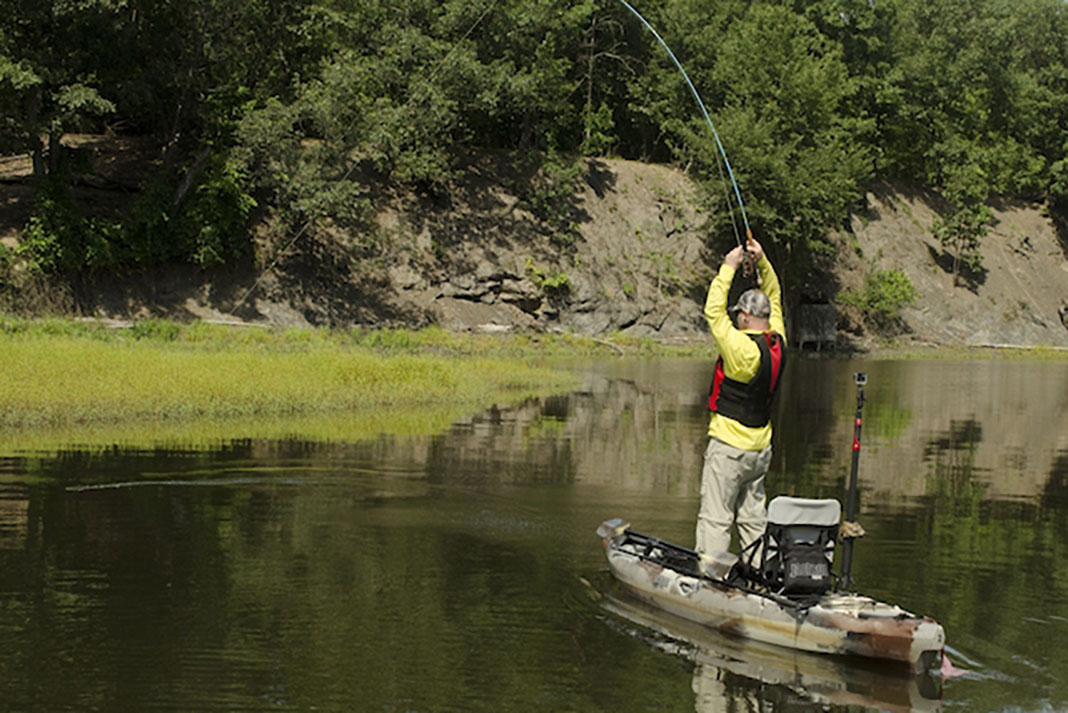 man stands and balances on his kayak without falling off while holding the fishing rod high overhead as a fish pulls drag