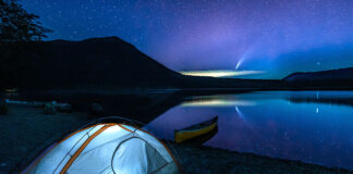 tent lit up at night with canoe beached on still lake and comet in sky