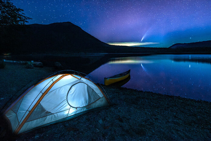 tent lit up at night with canoe beached on still lake and comet in sky