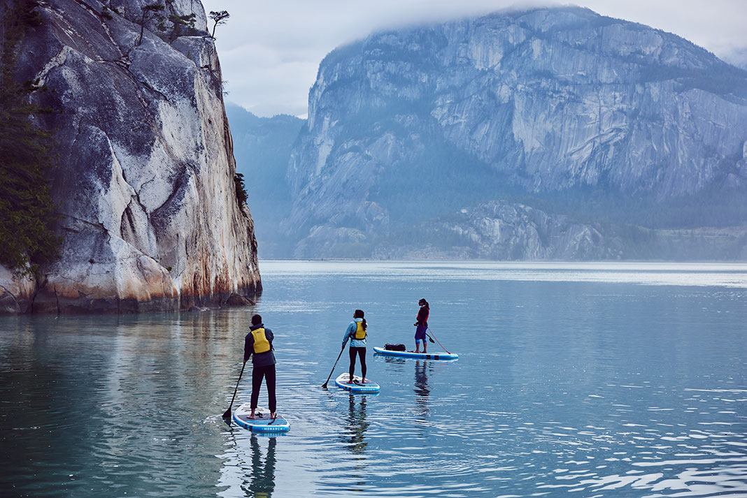 three people standup paddleboarding in front of towering cliffs near Squamish, BC