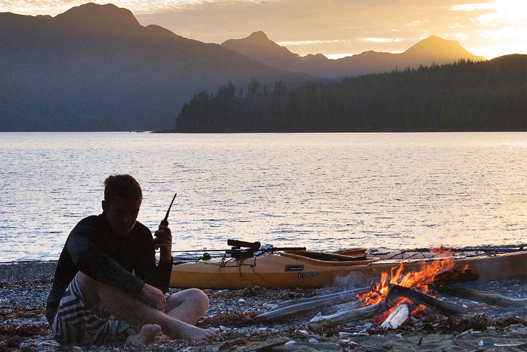 person sits beside campfire and beached kayak listening to radio for a weather prediction