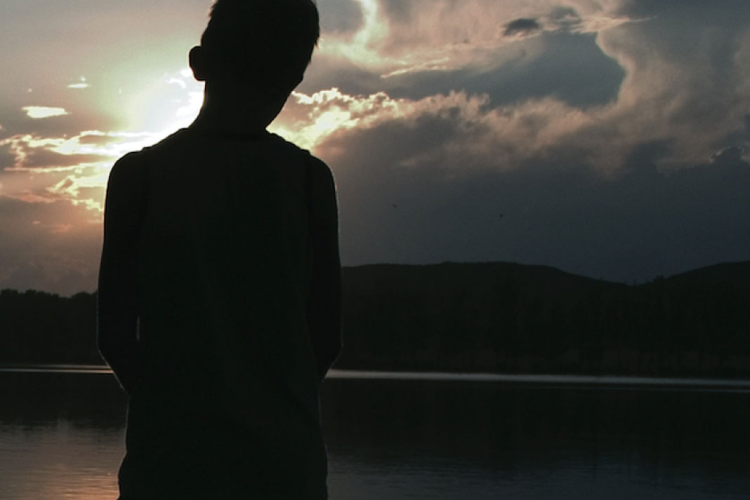 a boy stands silhouetted in front of a lake near dusk