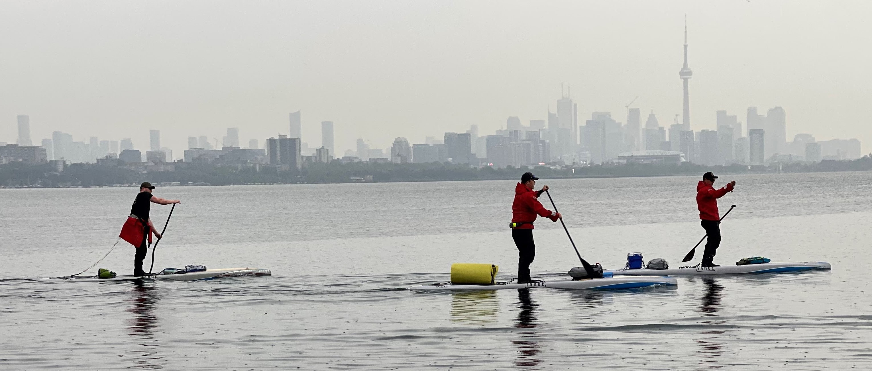 Three men on standup paddleboards with Toronto skyline in background.