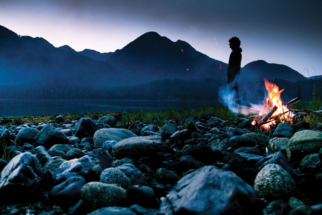 person stands on rocky beach with campfire