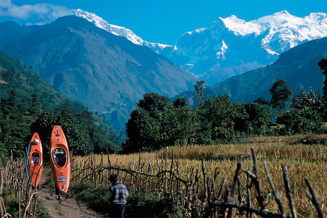two expedition kayakers carry their boats along a path through a field toward mountains