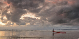 man stands at water's edge with a kayak in dramatic cloudy lighting
