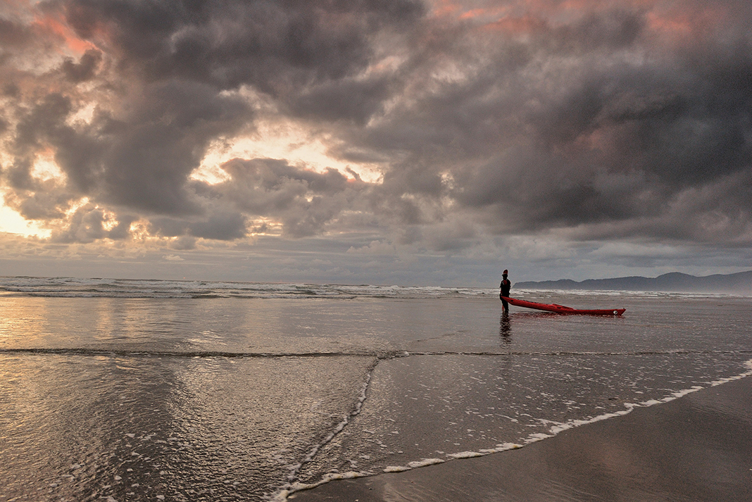 man stands at water's edge with a kayak in dramatic cloudy lighting