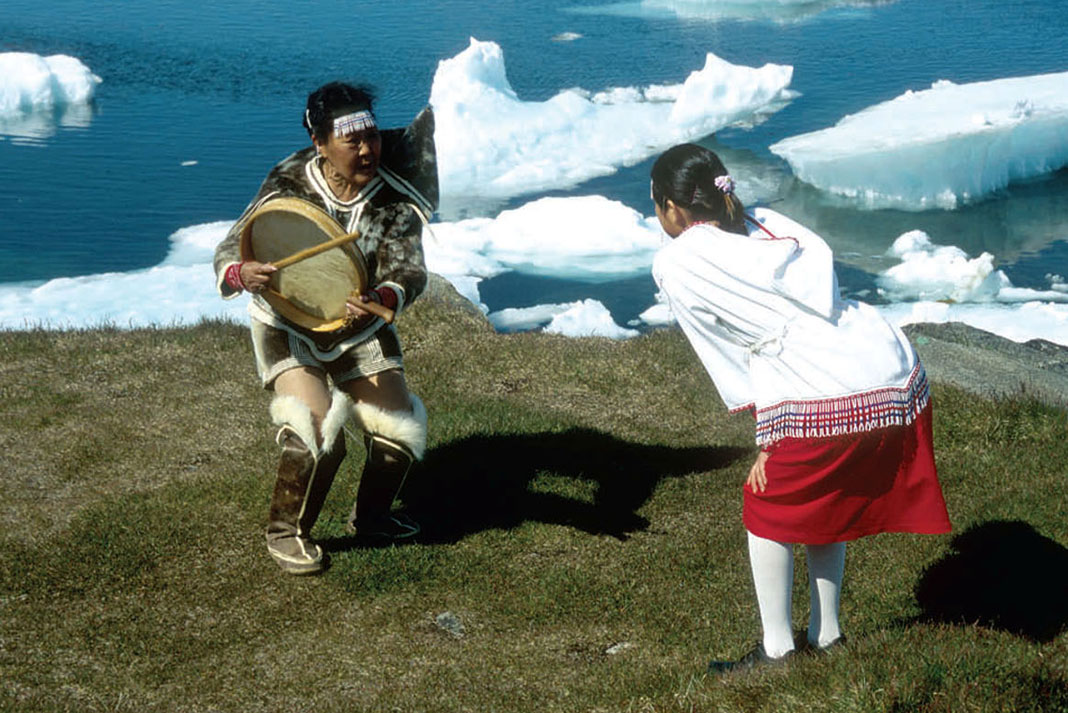 Two Greenland women practice drum dancing in front of a landscape of grass and icebergs
