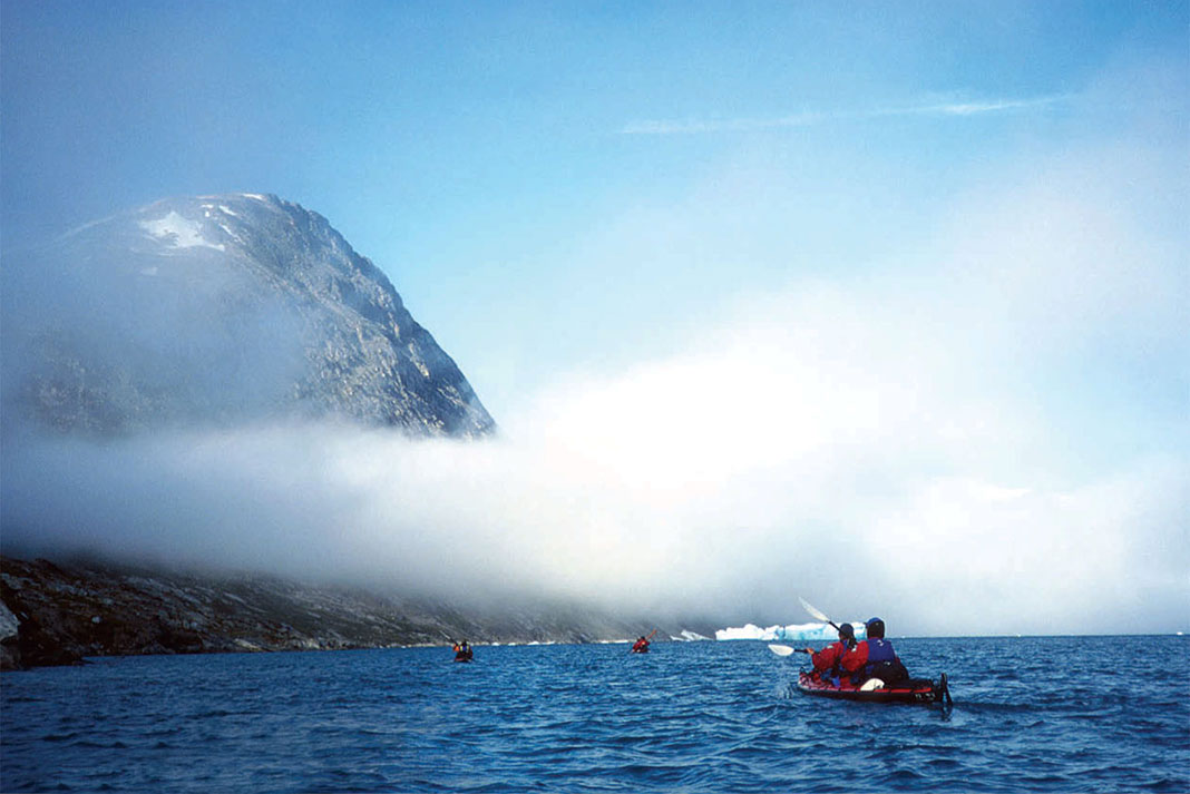 Tendrils of fog obscure the peaks of Ikateq Fjord as sea kayakers paddle in the foreground