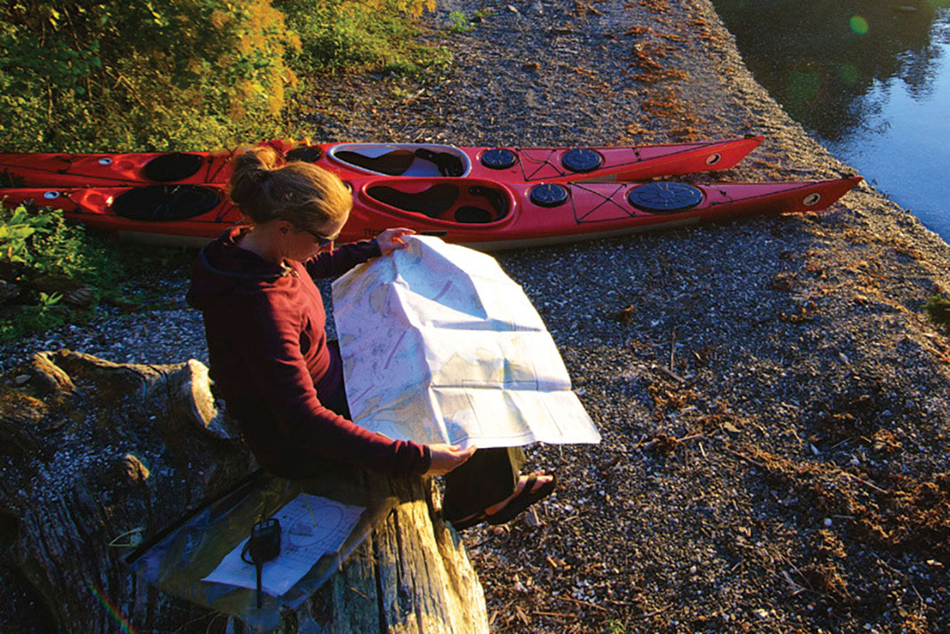 woman reads a map while sitting on a log at riverside beside her beached kayak