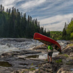 Magical Strokes: Celebrating The 100th Anniversary Of The Prospector Canoe person portages a Prospector canoe along the banks of a raging river