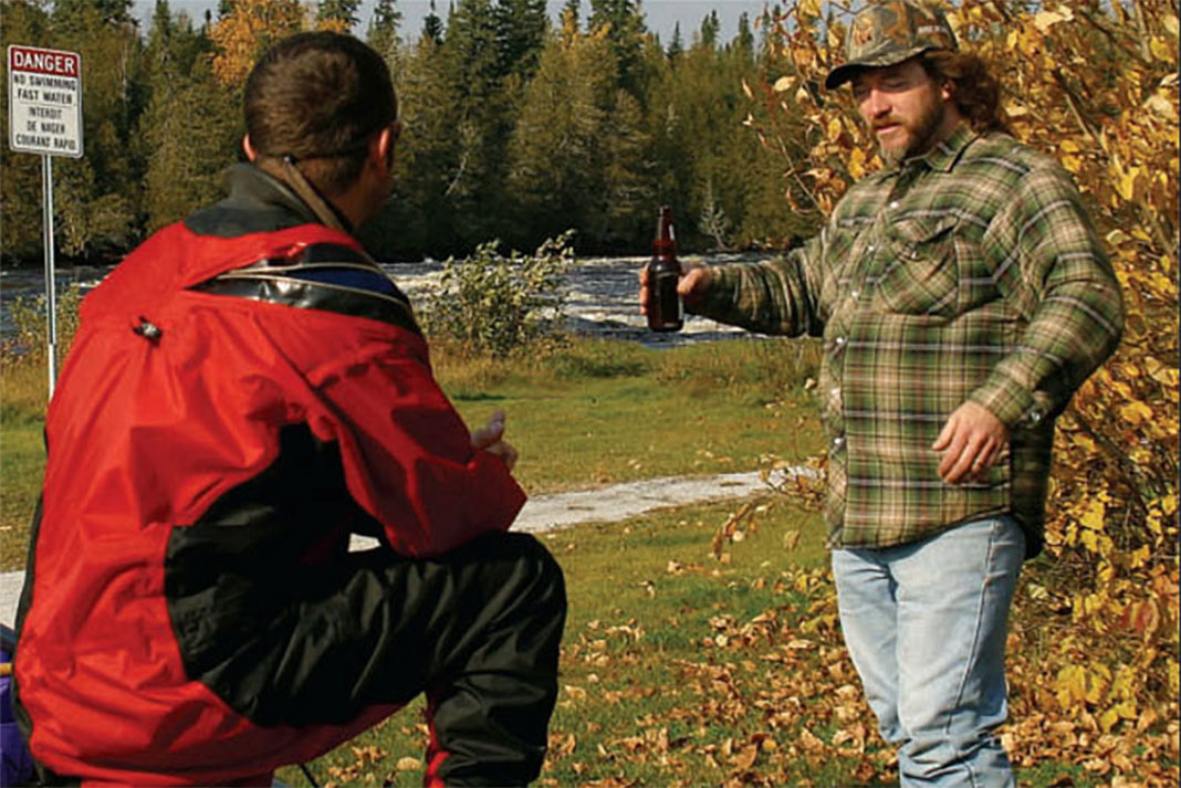 Man in whitewater paddling gear talks to man in hunting gear holding beer bottle