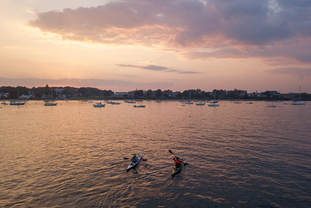 Man and woman paddling sea kayaks at sunset toward sailboats