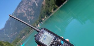 hang holding an ICOM M94D emergency communicator in front of a turqoise lake with kayakers paddling on it