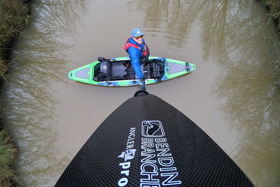 man holds up kayak fishing paddle to use as selfie stick while in kayak on muddy water