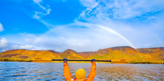 an autumn sea kayaker holds up their paddle with a rainbow in the background