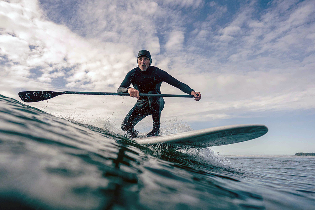 a man wearing a wetsuit surfs a wave on a hard paddleboard