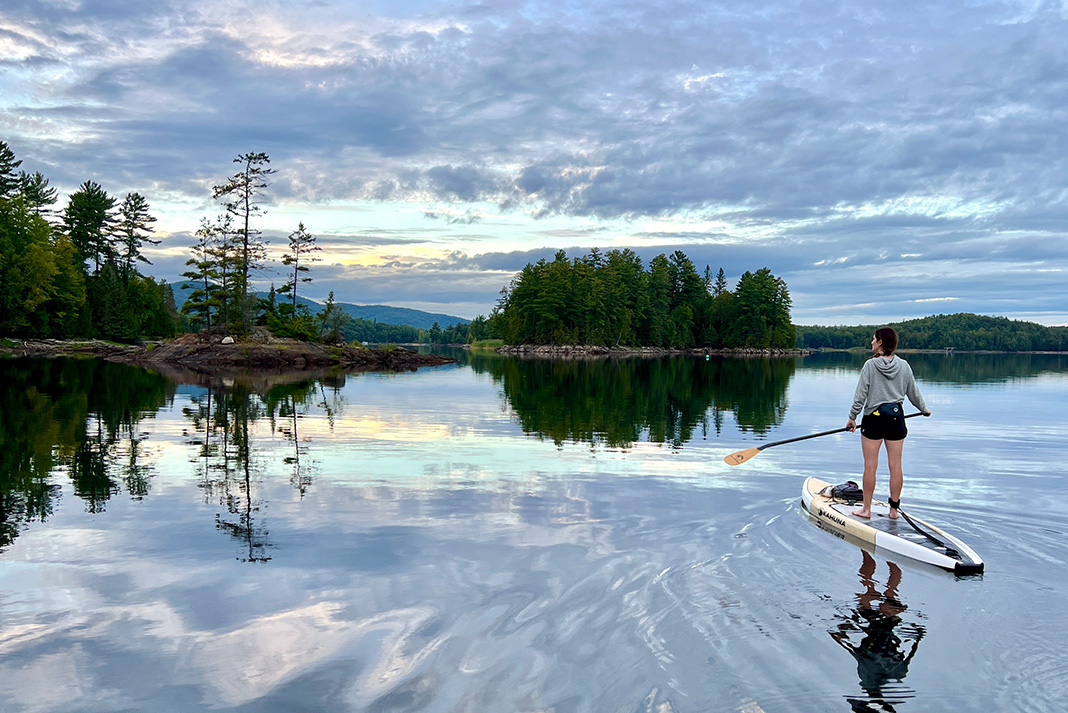 woman stands on a rigid paddleboard on calm water in front of scenic trees