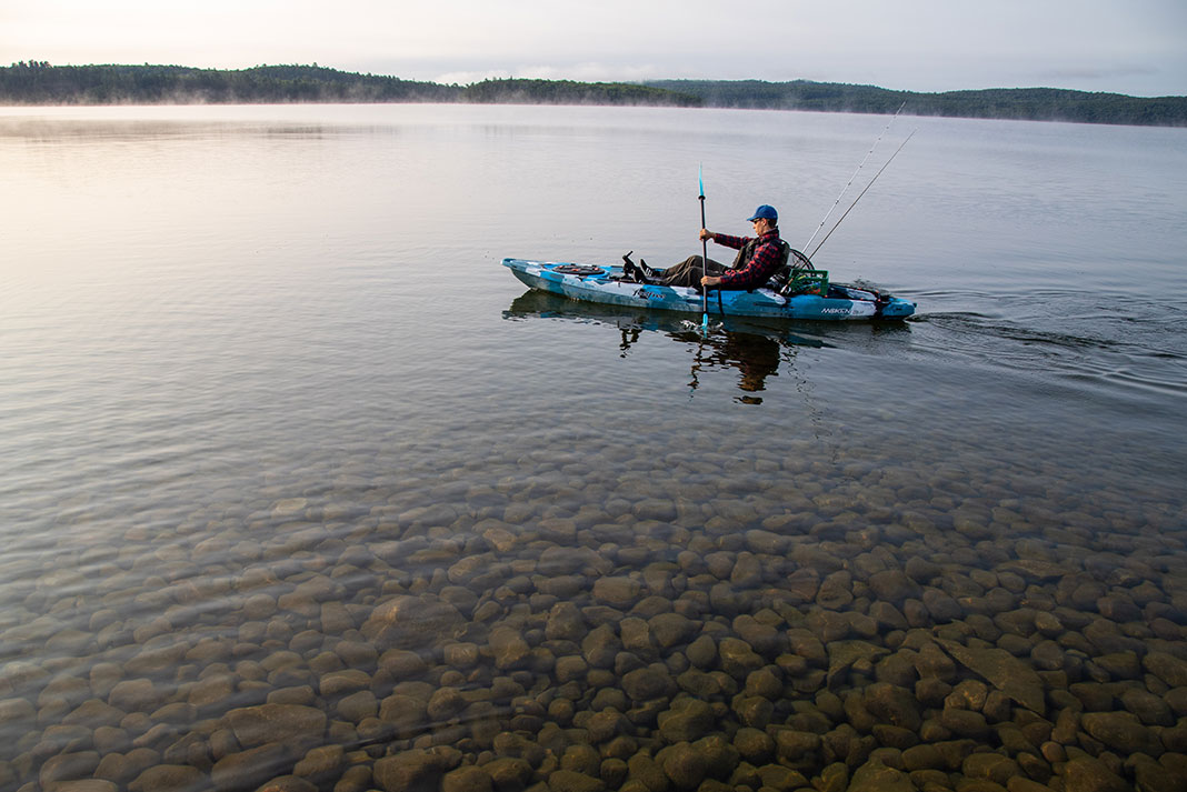 man paddling his kayak across calm waters with small rocks underneath