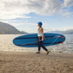 Below Board: Everything You Should Know About Paddleboard Fins woman carrying a paddleboard equipped with fins across a sandy beach