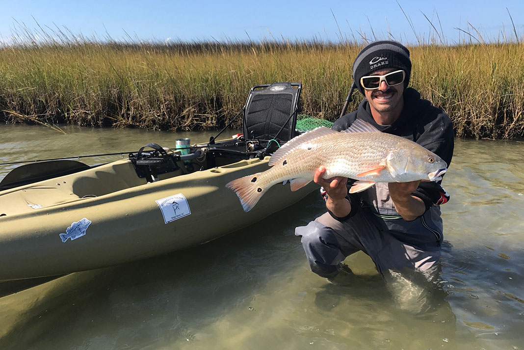 Man crouches in front of his fishing kayak holding a redfish caught in the flats