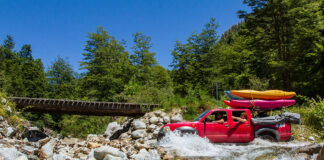paddlers in a shuttle rig drive across a rocky stream bed with a small wooden bridge in the background