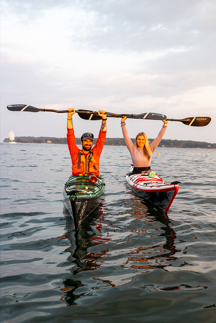 Man and woman in sea kayaks with paddles raised above their heads.