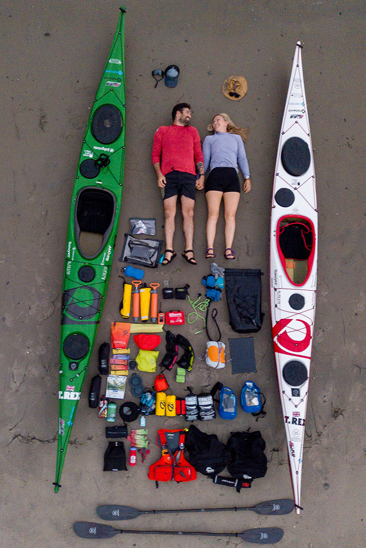 Woman and man lying between sea kayaks on a beach with gear spread out beneath them.