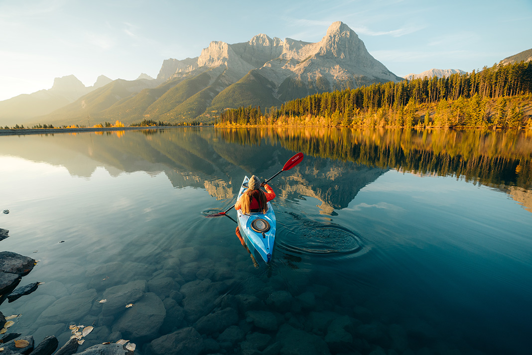 woman paddles kayak on still water in front of scenic mountains in golden light