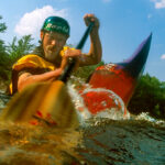 man paddling a whitewater boat and dipping his paddle into the water near the camera