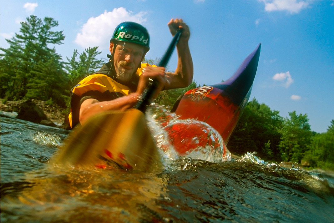man paddling a whitewater boat and dipping his paddle into the water near the camera