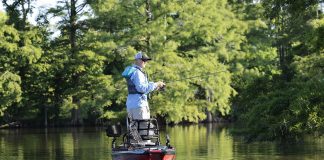 a man stands up and fishes from his kayak in front of a leafy backdrop