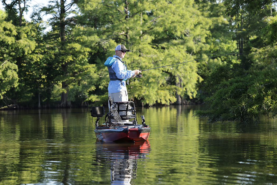 a man stands up and fishes from his kayak in front of a leafy backdrop