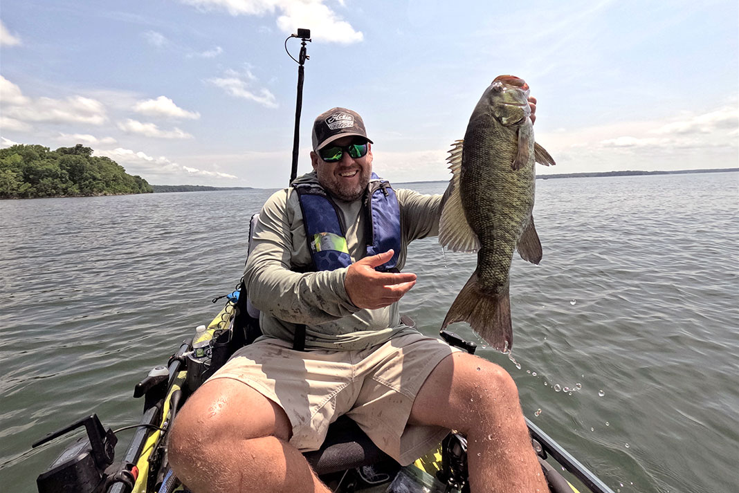 man smiles as he holds up a smallmouth caught on a sunny day