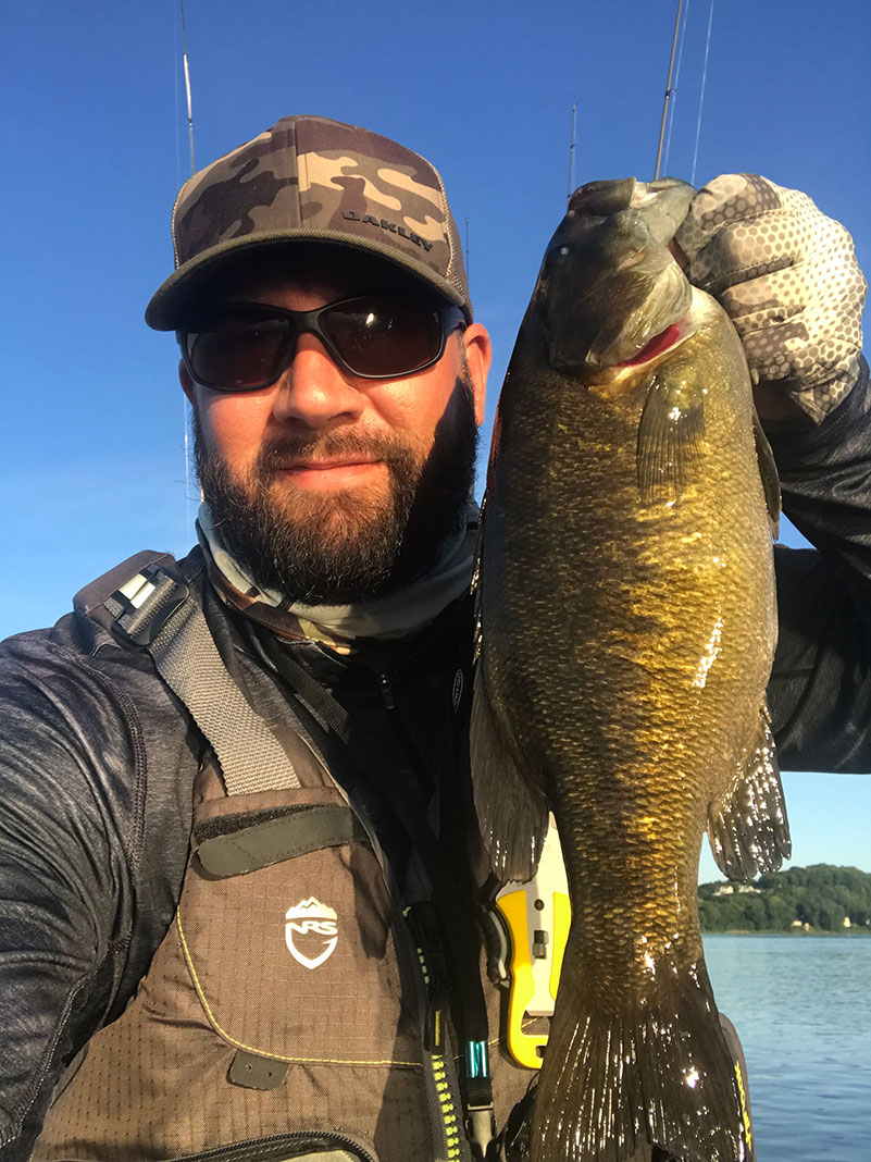 man wearing PFD, hat, gloves and sunglasses holds up a smallmouth