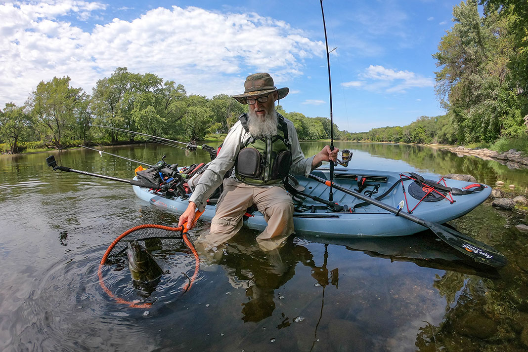 man with white beard uses net to land smallmouth bass by kayak