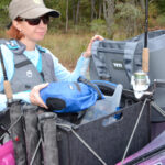 Woman standing beside fishing kayak with cooler, drybag, and other gear.