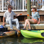 A couple has drinks on the dock by their fishing kayaks