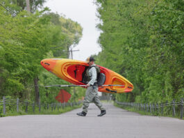 man walks across a narrow forest road while carrying an Old Town Vapor 10 kayak on his shoulder