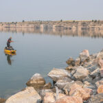 Find Out-Of-This-World, Untouched Fishing In Wyoming man standup fishing from a kayak in front of a rocky shore in Wyoming