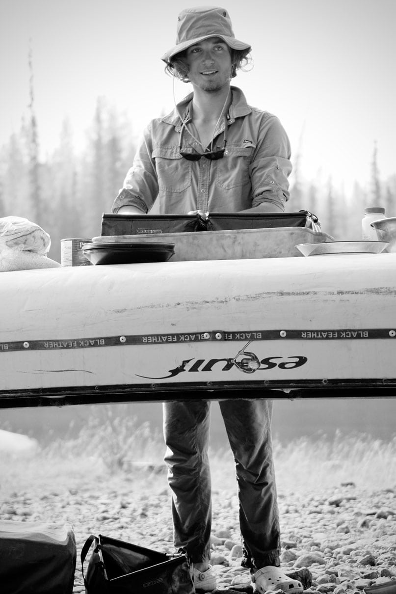 black & white photo of a young man in bucket hat washing dishes at a camp kitchen set up on an overturned canoe