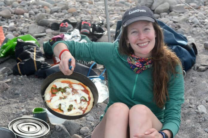 woman sitting near campfire holds up a cast iron skillet with backcountry pizza baked inside