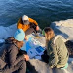 three women gather around a paper map spread out on rocks near the water, rather than using navigation apps