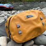 Best Waterproof Fanny Packs For 2026 The Patagonia Guidewater waterproof hip pack sitting on rocks near the edge of a river with a raft in the background.