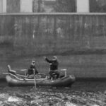 black and white photo of two men fishing from an inflatable boat beside large concrete bridge piers