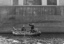 black and white photo of two men fishing from an inflatable boat beside large concrete bridge piers