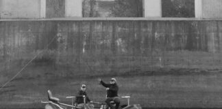 black and white photo of two men fishing from an inflatable boat beside large concrete bridge piers