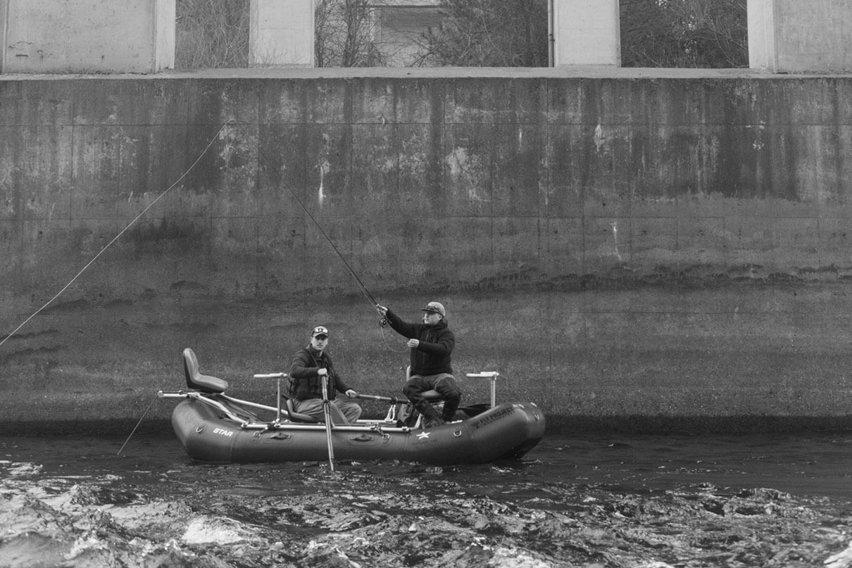black and white photo of two men fishing from an inflatable boat beside large concrete bridge piers
