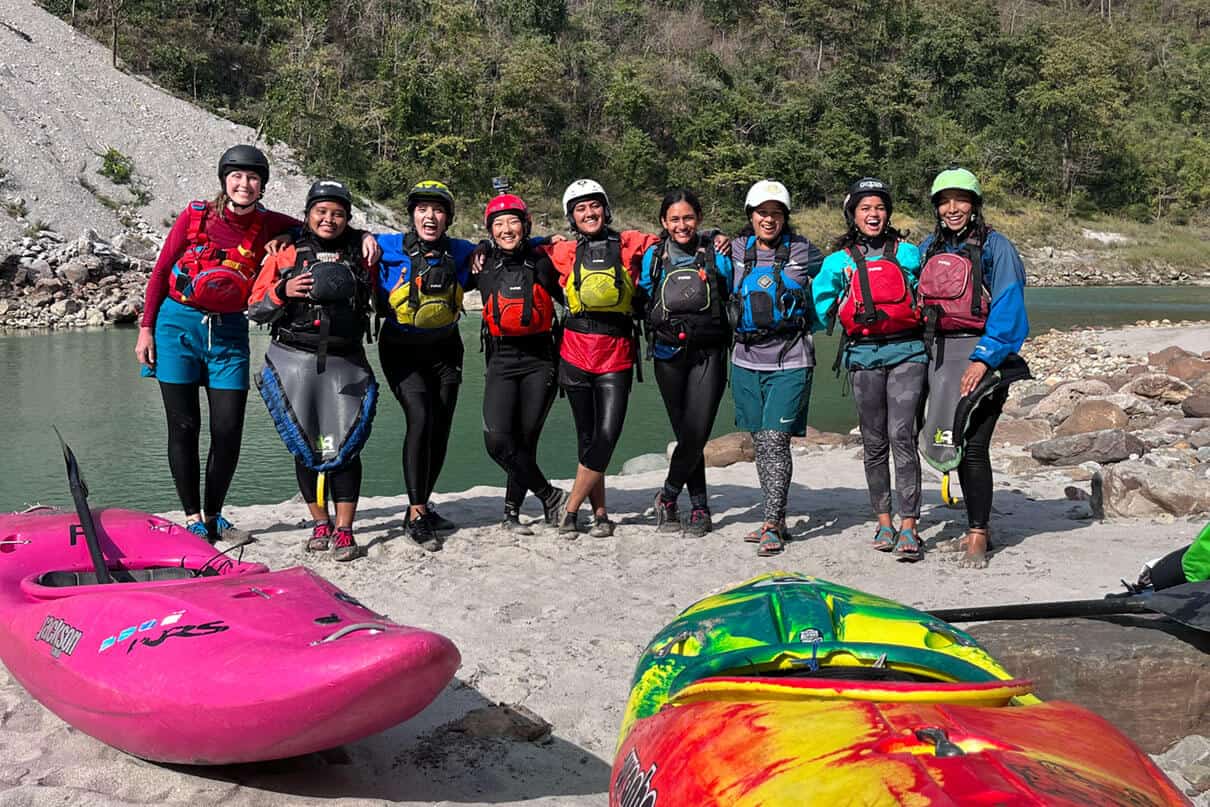 a group of woman kayakers pose for a group photo in their whitewater gear by a riverside in India